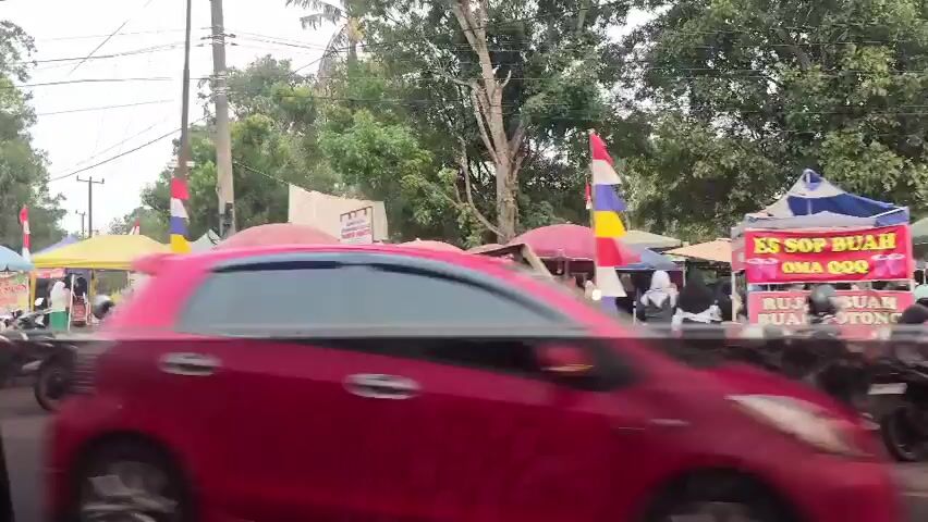 The video pans across an outdoor market. Numerous stalls are set up, some with red and white striped flags. Many motorcycles are parked in front of the stalls, and people are browsing. A red truck passes by on the left. The environment appears to be a semi-urban area with trees and overcast skies.