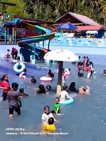 The video shows a crowded outdoor swimming pool. Many children are swimming and playing, some using inflatable rings. A colorful waterslide is visible in the background. There's a mushroom-shaped structure in the pool.  Trees and a building can be seen in the background. The video was recorded on an OPPO A6 Pro.