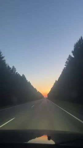 A first-person perspective video shows a car driving on a road bordered by trees on both sides. The sky is clear and blue, with the sun setting in the distance, casting an orange glow on the horizon. The headlights of the car are on, illuminating the road ahead. The reflection of the sunset is visible on the car's dashboard. The video is shot in portrait mode and appears to be taken at dusk.