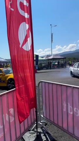 A yellow car with "KOMATSU TRUCK" written on the side drifts in a smoke-filled arena, followed closely by a white car, also drifting. Spectators stand behind a fence and in a raised seating area, watching the event. Mountains and a clear blue sky form the backdrop.