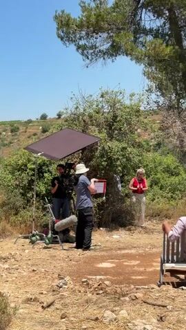 A film crew is set up outdoors. A woman in a white hat and blue shirt stands holding a red clipboard, facing away from the camera. To her left, a person operates a video camera on a tripod, holding a boom microphone. A large reflector umbrella is positioned above and behind the camera operator. To the right of the woman with the clipboard, a woman in a red top and light-colored pants stands with her hands clasped, facing a group of two people sitting on a bench in the foreground. The two people on the bench are viewed from behind; one has a brown hat and the other has dark hair with a headscarf. The environment appears to be a dry, rural area with trees and scrub brush under a clear blue sky.