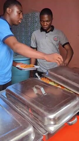 The video depicts an indoor scene in what appears to be a basic eatery or catering setup. Two men and one woman are present.

In the foreground, two men are engaged in serving food. The man on the left, wearing a blue short-sleeved polo shirt, is serving a portion of food, possibly rice, from a large metal chafing dish onto a white plate. He holds the plate with his left hand and uses a utensil to scoop the food. The man on the right, dressed in a grey short-sleeved polo shirt with a dark collar and a small circular logo on his left chest (featuring initials that appear to be "MT"), stands next to the serving dishes, observing the process. His hands are near the lids of the chafing dishes.

In the background, a woman wearing a blue polo shirt and a black head covering is seated on a white plastic chair, looking towards the activity. Behind her and to the right, two large blue barrels are visible, likely for water storage, and a rolled-up mattress with a blue and white patterned cover leans against the wall. The walls of the room are painted a light reddish-orange color. The scene suggests a communal meal service or food distribution.