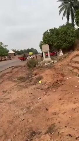 The video depicts a roadside scene in what appears to be a rural area. The ground is predominantly red dirt with scattered litter. A monument or structure stands near the side of the road. Several individuals are visible, along with various forms of transportation: a motorcycle-powered tricycle, a cart pulled by a white animal (likely an ox or cow), and a large yellow truck in the background. People are seen interacting with the animal and cart. The overall environment suggests a developing or agricultural area.