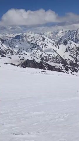 The video shows a panoramic view of snow-capped mountains under a blue sky with scattered clouds. In the foreground, there is a vast expanse of snow, with some tracks suggesting recent activity. Further back, there are some buildings and ski lift infrastructure, including towers and cables. The camera pans across the landscape, capturing the grandeur of the mountains and the snowy terrain.