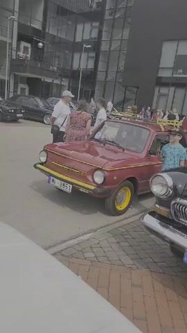 A gathering of people is observed around several vintage cars parked on a brick paved area in front of a modern building with large windows. The focus is on a red, four-door car with a yellow trim and a roof rack. A young boy in a blue patterned shirt is visible standing by the red car. To the right of the red car, a black vintage car with a prominent chrome grille and a blue light on its roof is parked. People are interacting with the cars and each other. The overall setting appears to be an outdoor event or car show.