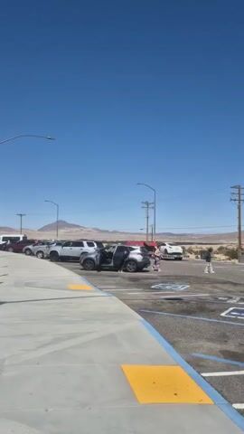 The video shows a parking lot in a desert environment. The sky is clear and blue. Mountains are visible in the distance. Several vehicles are parked in the lot, including cars and a Penske moving truck. Two people are walking through the parking lot. The parking spaces are marked with blue lines and handicap symbols. The ground is asphalt, and some yellow textured surfaces are visible near the sidewalk. There are also utility poles with wires overhead. The scene is bright and sunny.