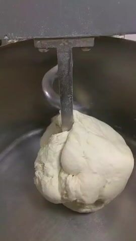 The video shows a dough hook kneading dough in a large, stainless steel mixing bowl. The dough is white and slightly sticky, and the hook is pulling and stretching it as it rotates. The bowl itself is clean and reflective. The mixer's arm is gray and sturdy, positioned above the bowl. The environment is likely a professional kitchen or bakery setting.