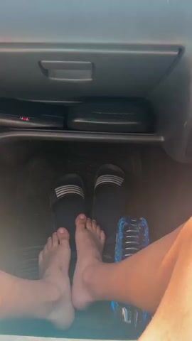 A person with bare feet inside a vehicle. The feet are resting near slippers in the driver’s footwell. The dashboard of the vehicle is in view, including the radio and vents. The car is moving on a highway with other cars visible through the windshield. The sky is blue and sunny.