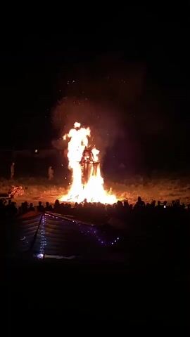 A large bonfire burns in a field at night. In the center of the fire is a wooden effigy that is also burning. A crowd of people are gathered in front of the bonfire, silhouetted against the flames. A tent with lights strung across it is in the foreground. The sky is dark, with some smoke rising from the fire. The scene appears festive and celebratory.