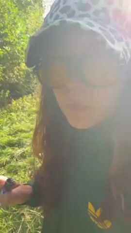 A person with long brown hair is smoking what appears to be a rolled cigarette or joint in a grassy field. The person wears large square sunglasses, a patterned bucket hat, and a green Adidas-branded tracksuit. They have a nose piercing. The lighting is bright, with the sun causing lens flare. The background shows a green field with trees in the distance. They hold a lighter in their hand.
