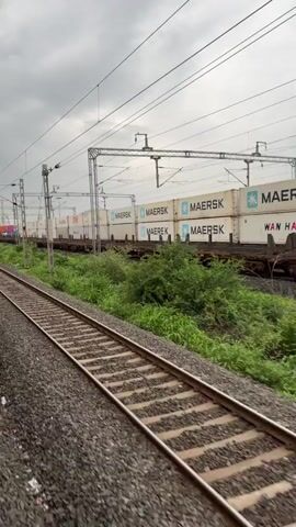 The video shows a train moving along a track. The train is carrying numerous shipping containers, many of which are branded with the "Maersk" logo. The train is traveling through a green, vegetated area with overhead electrical wires and support structures visible. The sky is overcast. The perspective is from another moving train traveling on an adjacent track, suggesting the video was filmed from a window.