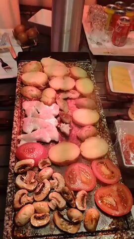 This is a close-up, top-down video of food being grilled on a rectangular hot plate. The hot plate is positioned on a wooden table outdoors, illuminated by a red outdoor heater. On the hot plate, there are several slices of potato, mushrooms, tomatoes, and some pieces of raw chicken. To the left of the hot plate, there are two plates with cutlery, and to the right, there are a few cans of soda and some glasses on a tray. There is also a small dish with chopped tomatoes. In the background, there's a roll of paper towels and some eggs in a bowl. The lighting is dim, with the red glow of the heater being the main light source. The video is static and does not show any people or active grilling.
