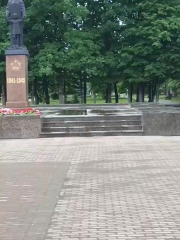The video shows a monument in a park. The monument is made of stone and has a statue of a man on top. There are also flowers around the base of the monument. The park has trees and a paved walkway. The video pans across the monument and then zooms in on the statue.