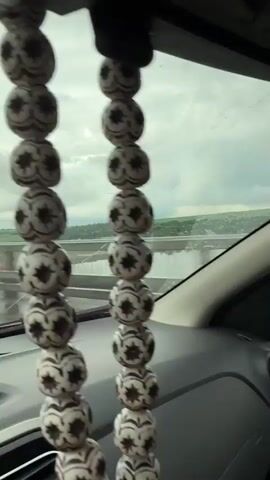 The video shows a view from the interior of a car moving across a bridge on a rainy day. The scenery includes the car's interior, the side window covered in raindrops, a side mirror showing the road, and a body of water with land in the distance beyond the bridge's railings. The sky is overcast.