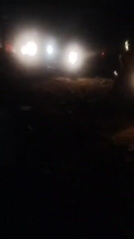 At night, bright headlights illuminate a dark, uneven dirt road. The lights belong to a vehicle that is partially visible on the right side of the frame. A building with a fence is dimly visible to the left of the lights. Debris litters the ground. The scene appears to be rural or industrial.