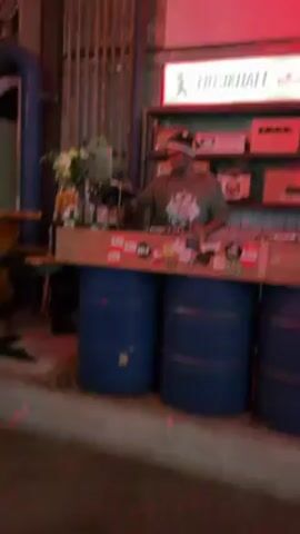 A medium shot shows a man DJing at a venue called "MUSIKHALL". The DJ, wearing a cap, glasses, and a graphic tee, is standing behind a makeshift DJ booth made of a wooden plank supported by three blue metal barrels.  The DJ has a laptop and other equipment on the surface. The front of the booth is covered in stickers. Behind the DJ is a shelf stocked with radios and other vintage electronics.  Red light illuminates the scene. The setting appears to be an indoor bar or event space. There are flowers and alcohol bottles on the DJ booth. The background includes a roll-up metal door, exposed pipes, and a partially visible projected screen. There are people near the frame's left side.