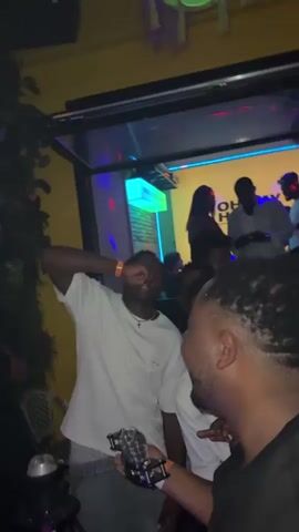 The video appears to be taken in a dimly lit indoor venue, possibly a bar or club. The primary focus is on a group of people, predominantly young adults. In the foreground, a person wearing a white t-shirt is taking a shot. Another person with sunglasses and a black t-shirt is holding a drink. Several other individuals are visible in the background, some dancing and interacting. The environment features yellow walls, neon lighting, and foliage decorations. The overall atmosphere seems lively and celebratory.