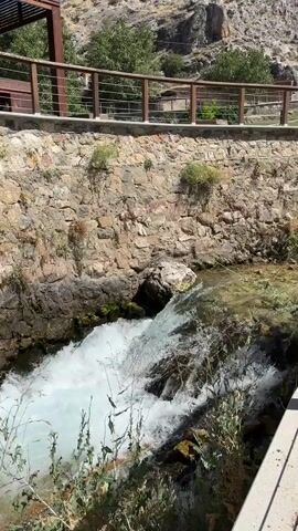 The video shows a fast-flowing stream of water rushing through a narrow channel. The water is white and frothy, indicating a strong current. The channel is lined with rough stone walls, with some vegetation growing on top of the walls and along the banks. In the background, there is a rocky hillside with some trees. A wooden railing with metal posts runs along the top of the stone wall, suggesting a walkway or viewing area above the stream. The light suggests it is daytime.