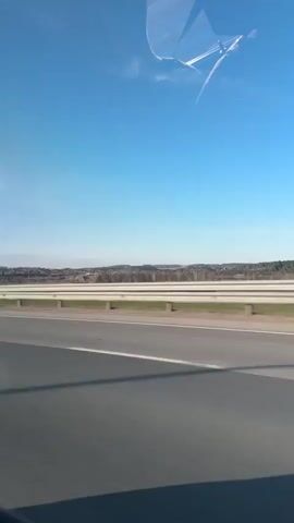 The video shows the inside of a car moving on a highway, as seen through the passenger's window and from the backseat. Initially, the road, a barrier, and a distant landscape are visible. A large truck passes in the adjacent lane. The perspective shifts to the car's interior, showing the safety belt and the head of a passenger in the front seat. The focus remains on the passing scenery through the window. The sky is bright blue. The car appears to be moving at a consistent speed.