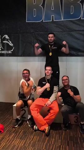A group of four men are posing for a photo on a stage. Three men are sitting on podiums labeled 1, 2, and 3. A fourth man is standing behind them. All four men are wearing medals around their necks. The man on podium 2 is wearing orange pants and has his arms crossed. The man on podium 1 is wearing glasses and flexing his arm. The man on podium 3 is also wearing glasses and smiling. The man standing behind them is wearing a black t-shirt and flexing his arms. There is a banner behind them with text that is partially obscured but appears to say "BAB" and "WRESTLING". The floor is made of wood planks.
