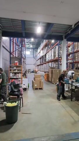 The video depicts an active industrial warehouse or fulfillment center located in Molokovo, Russia. The scene is dominated by tall, industrial shelving racks fully stocked with numerous packaged goods, indicating a significant storage capacity. In the foreground and middle ground, several workers, mostly men dressed in work attire such as overalls and hoodies, are engaged in packaging tasks at a line of tables.

The primary activity involves workers handling distinct teal-colored product boxes. They appear to be preparing these items for shipment, possibly by packaging them into larger cardboard boxes or applying protective wrapping. One worker walks into the frame carrying a teal box, places it on a table, and joins his colleagues in the packing process. The consistent sounds of packaging tape being pulled and used are audible throughout the video, emphasizing the ongoing work.

The warehouse environment features a concrete floor and is brightly illuminated by artificial overhead lights. Various objects are visible, including cardboard boxes on pallets and shelves, rolls of packaging film or wrap, and a speaker on the floor near a workstation. The overall impression is one of a busy and efficient logistics operation, with workers focused on their tasks. No specific text is clearly visible on any of the packages or signage.