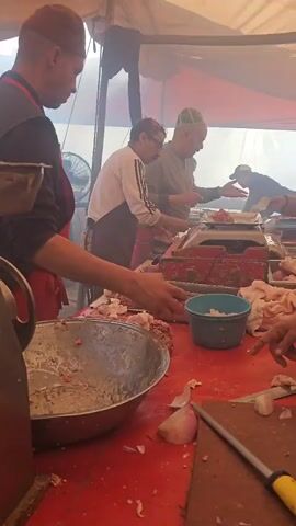 A group of people work together processing meat under a large tent. Several men are preparing raw meat, possibly for sale or consumption. They are weighing meat on a scale and chopping ingredients like onions on a red countertop. There are metal bowls filled with chopped meat and a smaller blue bowl. Behind the workers, other people are seated at red tables drinking tea and conversing, appearing to be customers or patrons. The tent is filled with a smoky haze.