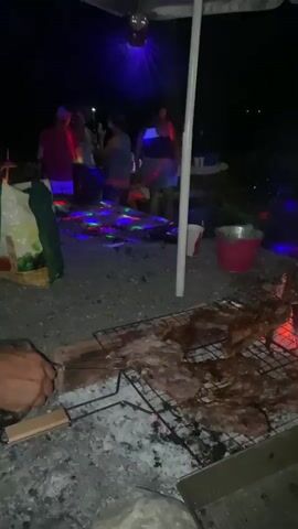 This is a nighttime video of an outdoor gathering. In the foreground, there is a grill with meat cooking over coals. A man in a white t-shirt and red shorts is tending to the grill. In the background, several people are dancing under disco lights and an umbrella. There are also bags and buckets visible. The overall atmosphere appears festive.