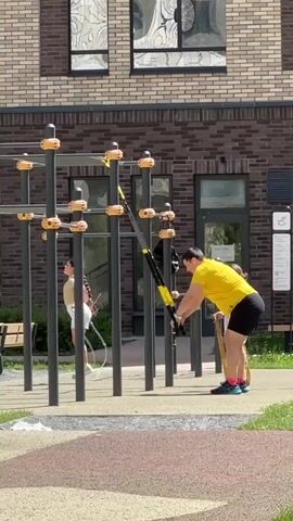 A man in a yellow t-shirt and black shorts is performing an exercise using a TRX suspension trainer on an outdoor sports ground. He is holding onto the straps with his arms extended, leaning back with his body at an angle. A young girl in a light-colored top and white shorts is standing to the left, also near the exercise equipment, facing away from the camera. She appears to be looking towards the man. The sports ground has a textured surface, with areas of pink and grey, and is bordered by grass. Behind the exercise equipment, there is a modern brick building with large windows. The scene is set during the daytime, with bright sunlight casting shadows. The overall atmosphere suggests an active, recreational setting. The building has visible windows with intricate patterns reflected in them. There is a sign on the building that appears to have text and an icon, but it is not legible in detail. The time of day appears to be sunny and bright, suggesting a warm day.
