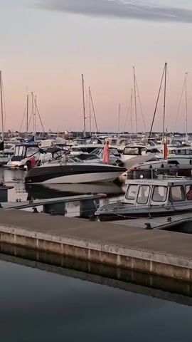 A marina at sunset with several boats docked. A medium-sized, white and black speedboat is moving slowly through the water. Two people are on board the speedboat. One person, wearing a pink dress, is standing near the stern. Another person, wearing a light-colored shirt and dark pants, is standing near the helm. A smaller, silver boat is docked in the foreground. The sky is a soft pink and orange, and the water is calm. Sailboats with tall masts are visible in the background.