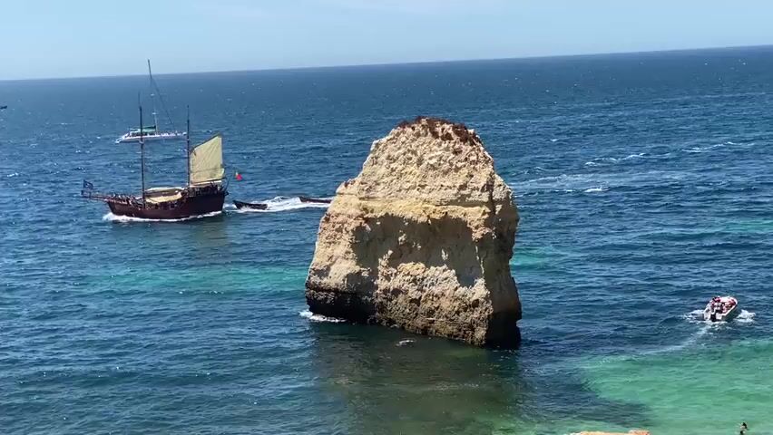 The video shows a scenic view of the ocean on a sunny day. In the foreground, a large rock formation stands tall in the clear blue water. To the left, a wooden sailboat with a single sail is moving through the water, leaving a white wake. Behind the sailboat, a smaller white boat is also visible. In the distance, the blue ocean stretches out to the horizon under a clear sky. The water appears to be a mix of deep blue and turquoise colors, suggesting shallow areas near the shore. The overall scene conveys a sense of calm and natural beauty.