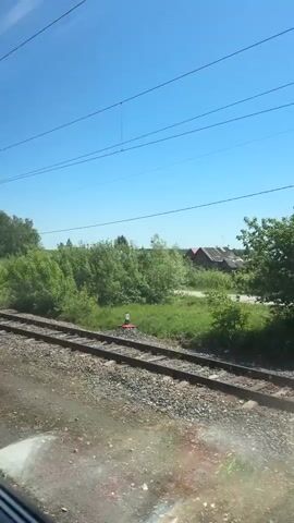 The video shows a view from a moving train. The train tracks run alongside a grassy area, followed by a row of green bushes and trees. Beyond the greenery, there are several houses with varying roof colors. Overhead, power lines and a railway electrification pole stretch across the bright blue sky. The gravel area between the viewer and the train tracks has a wet, reflective surface.