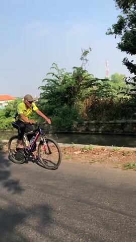 A person is riding a bicycle on an asphalt road. The person is wearing a yellow shirt with green and black accents, black pants with white stripes, a green baseball cap, sunglasses, and flip-flops. The bicycle is a mountain bike with a dark frame and multi-colored accents. The person is pedaling and appears to be moving forward. The environment is outdoors with trees and a building visible in the background. There is a curb and a ditch or water feature alongside the road. The lighting suggests it is daytime.