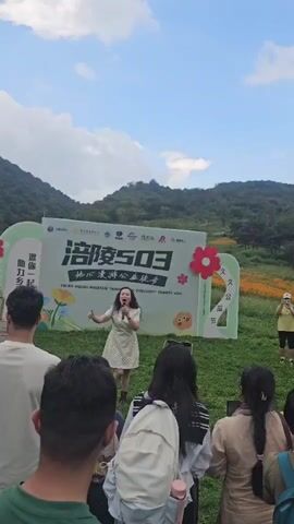 A woman in a polka-dot dress sings into a microphone on a grassy stage, with a large banner behind her that reads "PULING MOUNTAIN INNER EARTH DISCOVERY CHARITY HIKE." The banner features Chinese characters, logos, and cartoon flowers. A crowd of people, mostly seen from the back, watches her perform. In the background, a lush green mountain is visible under a cloudy sky. The woman is smiling and gesturing with her free hand, appearing to engage with the audience. The overall atmosphere seems festive and community-oriented, possibly marking the start or end of a charity event.