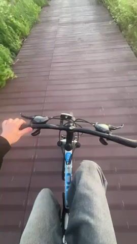 A first-person perspective video shows a person riding a bicycle on a wooden path. The path is lined with green grass on either side. In the distance, the sky is a gradient of blue, orange, and yellow, indicating either sunrise or sunset. The sun is visible on the horizon. The person's legs, wearing jeans, and hands on the handlebars are visible in the foreground. The bicycle is blue and white. The video appears to be shot outdoors during daytime.