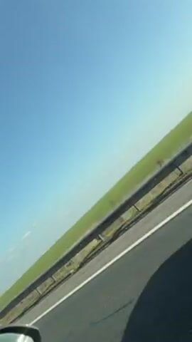 The video appears to be taken from inside a moving vehicle on a highway. The initial shot shows a highway sign indicating the directions to Constanta and Fetesti, with a green field in the background under a clear blue sky. The camera then shifts to capture a large yellow shipping container being transported on the back of a truck moving in the same direction. The container is secured with multiple straps and has caution signs. The view then returns to the scenery alongside the highway. The predominant colors are blue, green, yellow, and grey.