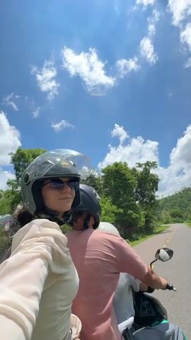 A first-person perspective video shows three people riding on a scooter on a paved road surrounded by lush green trees and hills under a bright blue sky with scattered white clouds. The person in the foreground, on the left, is wearing a light-colored sheer top, sunglasses, and a grey helmet. They are looking to their right. Behind them, another person is wearing a pink shirt and a black helmet. The third person, wearing a white helmet, is seated behind the person in the pink shirt. The person in the foreground briefly flashes a peace sign at the camera. The scooter is moving forward, and the scenery is passing by.