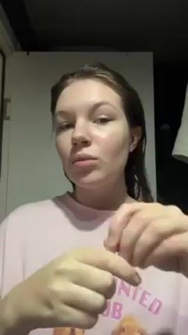 A person is standing indoors in a bathroom. The person is fair-skinned with light brown hair that is wet, wearing a light pink t-shirt with cartoon characters. The person opens a small glass vial, pours the contents onto their hand, and rubs it into their face. The person is looking at the camera during the clip. A bathroom door and a white towel are visible in the background.