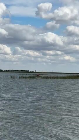 The video shows a wide shot of a body of water under a cloudy blue sky. In the distance, a thin strip of land with tall grass or reeds is visible, and on the right side of the frame, there appears to be a person in an orange shirt wading in the shallow water near the reeds. The water is choppy, with small waves. The overall scene is outdoors and appears to be a natural, possibly marshy or lake environment.
