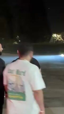 The video shows a group of people watching a black car doing donuts in an empty lot at night. The car is spinning around in a circle, kicking up smoke. Some people are standing close to the car, while others are farther away. There are buildings and trees visible in the background.
