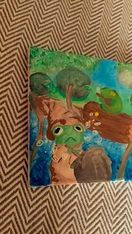 The video pans over a painting of frogs in a pond. The painting depicts several green frogs, some appearing to be in water with lily pads. The artwork is displayed on a textured surface, possibly a rug or blanket with a chevron pattern. The lighting suggests it is indoors.