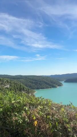 A vertical video captures a scenic overlook of a turquoise lake bordered by lush green forested hills under a bright blue sky with scattered clouds. The foreground is filled with the tops of green bushes and some dry branches, partially obscuring the view of the lake and hills. The sun is visible high in the sky, casting a glare. The camera appears to be stationary.