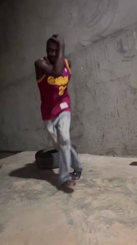 A dark-skinned man stands in an enclosed space with concrete walls and floor. He is wearing a maroon basketball jersey with the number "23" in yellow and light-colored denim jeans. He is also wearing dark-colored sandals. A used car tire is on the floor behind him. The man is looking up and adjusting his jeans. He appears to be standing still. A blue metallic structure is visible in the background.