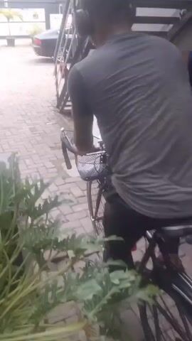A person wearing headphones and a grey t-shirt rides a bicycle down a paved walkway. The bicycle has a basket on the handlebars and a red reflector on the rear fender. To the left of the walkway are large green plants and a grey car. To the right are metal stairs leading up to a building. In the background, there are more buildings and a gate. The sky is overcast.