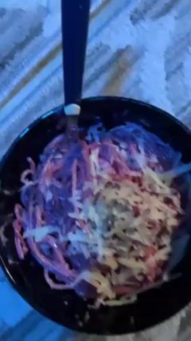 A man sits shirtless on a blue carpet, eating pink and purple pasta with shredded cheese from a black bowl with a red plastic fork. He has tattoos on his arms and an earring. He looks to the side as he eats. The background is cluttered with items, including a black duffel bag.