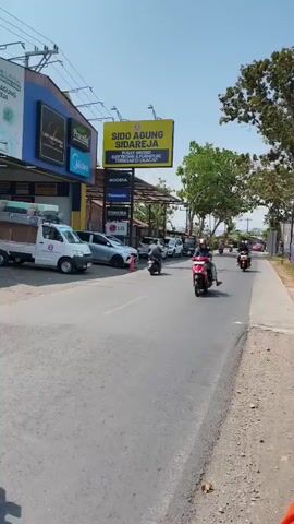 A still shot captures a bright, sunny day on a paved road with a sidewalk on the right and businesses on the left. A billboard for SIDO AGUNG SIDAREJA, advertising electrical and furniture items, stands prominently on the left side of the road. Several cars are parked in front of the businesses. Two motorcycles are on the road. The first motorcycle, ridden by a person wearing a helmet and a blue jacket, is in the foreground, moving away from the camera. The second motorcycle, with two occupants, is further down the road, also moving away. Trees line the right side of the road, with some red and white decorations visible. The sky is clear and blue.