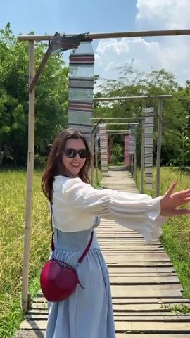 A woman is walking away from the camera on a wooden bridge. She is wearing a light blue dress, a white cardigan, and sunglasses, and carrying a red heart-shaped purse. The bridge is surrounded by tall grass and trees and decorated with hanging rectangular fabric. The sky is blue with white clouds. The woman waves as she walks away.