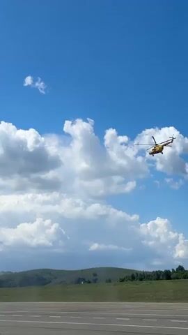 A helicopter is visible in the sky above a rural landscape. The helicopter is yellow and red and is flying among white clouds. The ground below is green and hilly, with a road or runway in the foreground. The sky is blue.