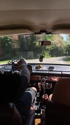 The video shows a person driving a car from the perspective of the backseat. The driver, who is bald and has tattoos, is wearing a black shirt and ripped jeans. They are holding a steering wheel that is decorated with a chain. In the passenger seat, another person's legs are visible. The car's interior is visible, including the dashboard with several gauges and a gear shift. Hanging from the rearview mirror are two small ornaments, one resembling a green frog and the other a yellow pom-pom. The car is driving on a paved road surrounded by trees and foliage. The sun is shining brightly, casting shadows inside the car. The driver appears to be talking or reacting to something, based on their facial expression and hand gestures.