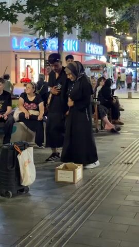 A group of people are standing on a sidewalk in front of a store. A man and a woman in a hijab are looking at a phone. A donation box is on the ground in front of them. There are other people sitting on a bench and standing around. The store behind them has a large sign that says "LC Waikiki". The lighting suggests it is evening.