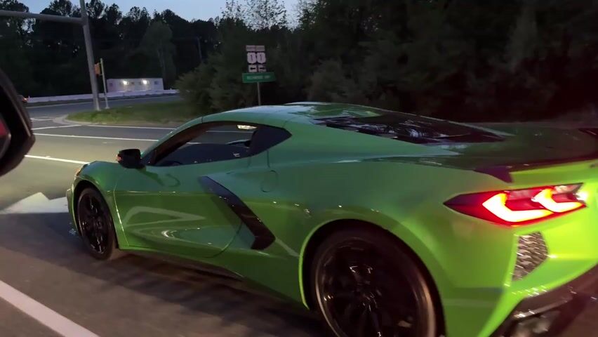 The video shows a vibrant green Chevrolet Corvette C8 accelerating away from a stoplight at dusk. The camera is positioned inside another car, looking out the driver's side. As the Corvette moves forward, it becomes fully visible, showcasing its sleek design and black rims. The stoplight transitions to green, and other vehicles start moving as well. The background includes trees and buildings with illuminated lights.
